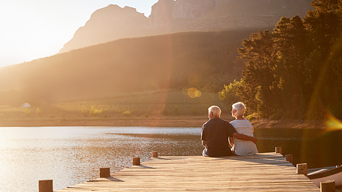 Ruhestand – Achtung, Stolperfalle! Sitzendes Rentnerpaar am Steg mit Blick auf den See.