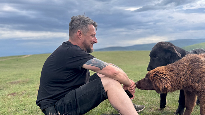 Auf den Hund gekommen Gerd Schuster sitzt auf einer Wiese, zwei Hunde sitzen vor ihm. | © privat